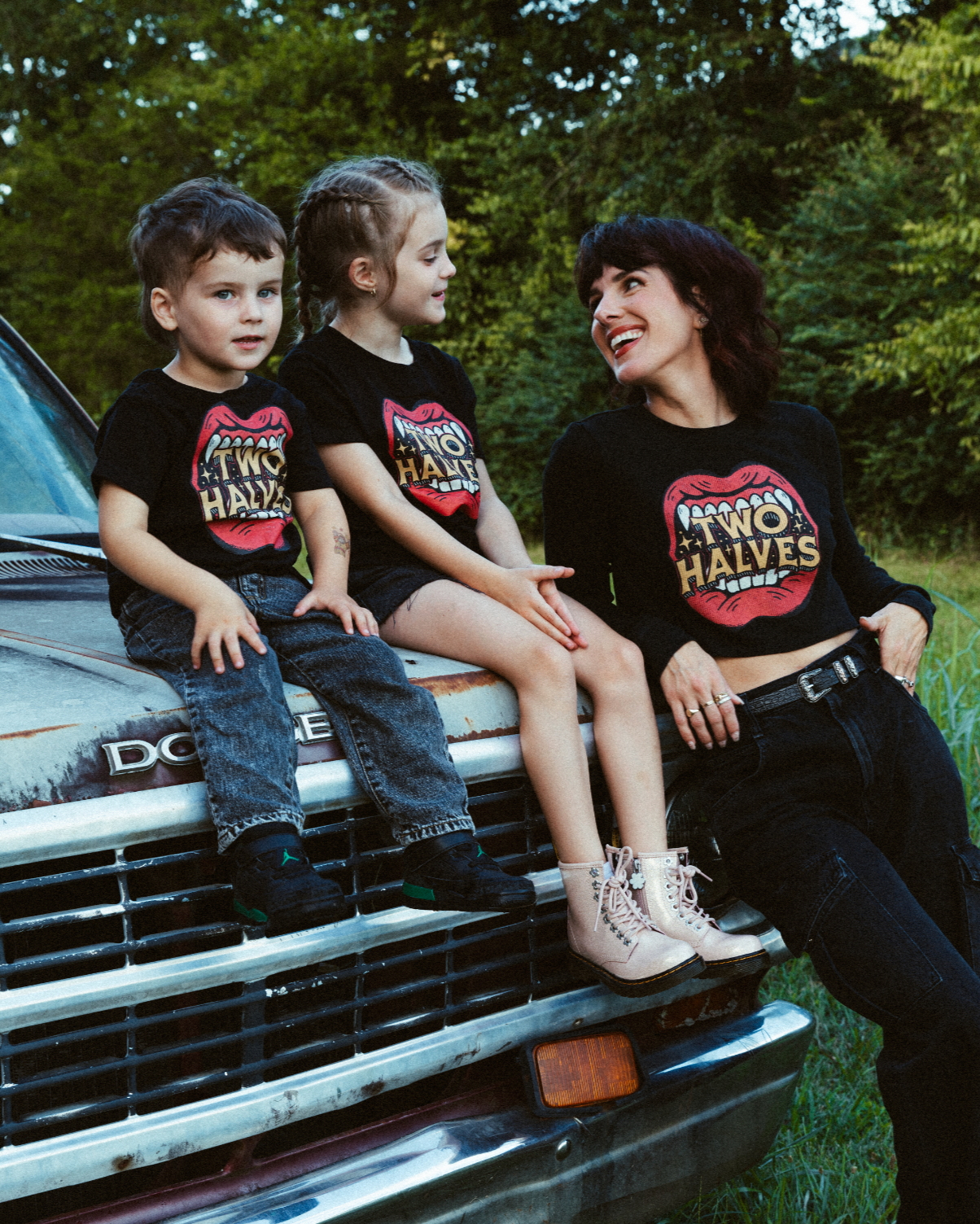 Three people sitting on a truck with 'Two Halves' shirts in a natural setting